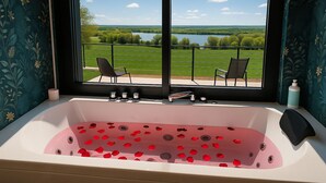 A bathroom with a bathtub filled with rose petals, a window with a view of a green landscape and a lake, and two chairs on an outdoor balcony. - Maison du lac, 1h from Lyon and Genèves, Jacuzzy, swimming pools, magnificent lake view. (Jayat)