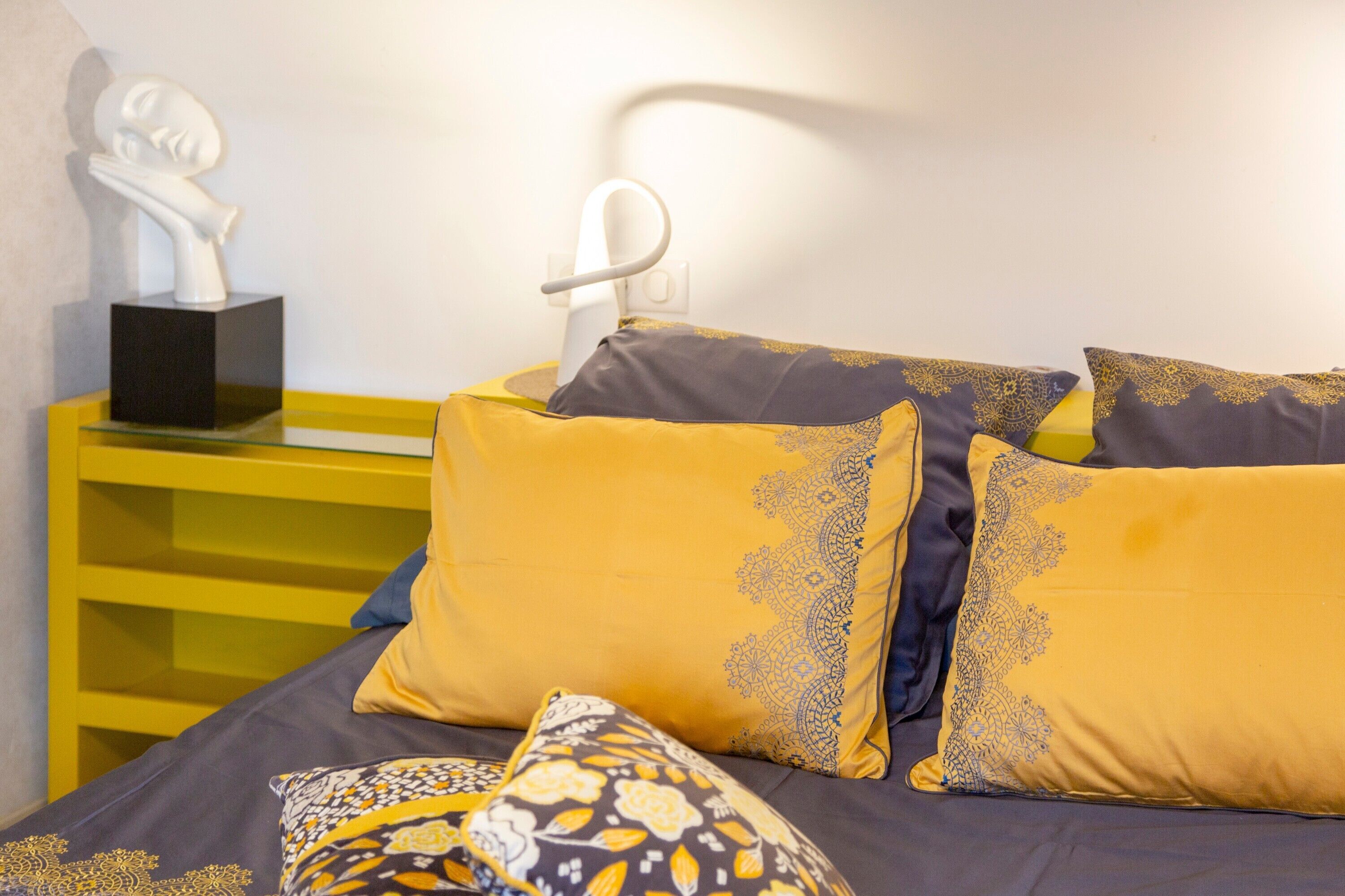 Una cama con almohadones estampados, una escultura blanca sobre un pedestal negro y una mesita de noche amarilla.