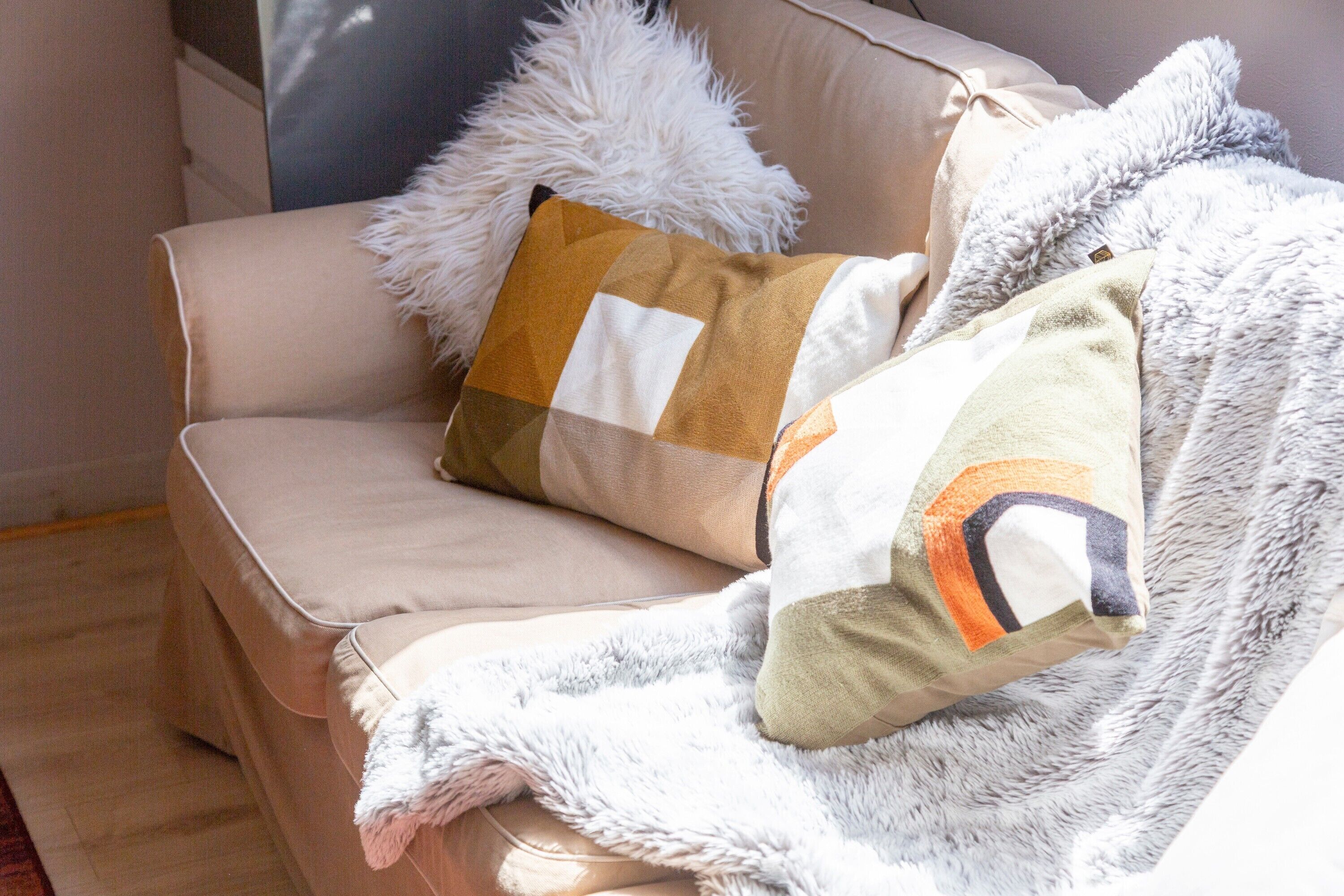 A beige sofa with a fluffy white pillow and a patterned pillow.