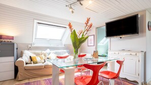 A modern living room with a glass dining table, red chairs, a beige sofa, a flat-screen TV, and a large window. - Holiday Home 'Les Géraniums À 800m Plage' with Private Terrace, Shared Garden and Wi-Fi (Kerlaz)