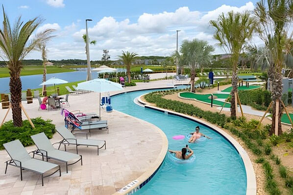 A swimming pool with lounge chairs and umbrellas, surrounded by palm trees and a golf course in the background.