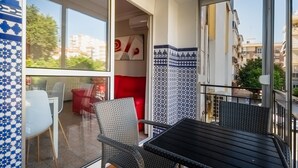 Een balkon met rieten stoelen en een tafel, een rode stoel en uitzicht op gebouwen en bomen.
