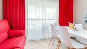 Een kamer met een rode bank, een witte eettafel met stoelen en een raam met dunne gordijnen.