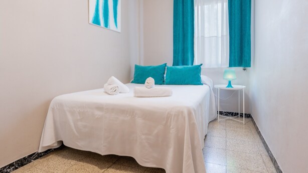 Hotelkamer met een bed, blauwgroene gordijnen, een witte spreidek, een wit kussen, een witte handdoek, een kleine tafel met een lamp, en een lijst met een afbeelding aan de muur.
