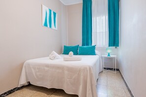 Habitación de hotel con cama, cortinas azul verdosas, colcha blanca, almohada blanca, toalla blanca, una mesita con lámpara y un cuadro en la pared.