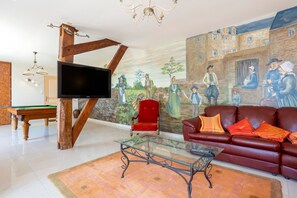 Een woonkamer met een biljarttafel, een rode stoel, een glazen salontafel, een leren bank en een muurschildering van historische figuren.