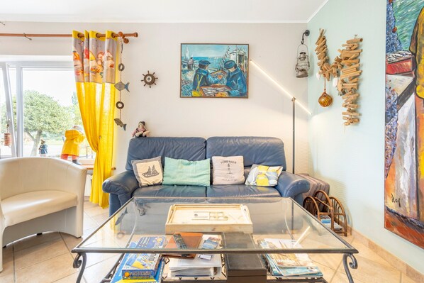 Uma sala de estar com uma mesa de centro de vidro, um sofá azul e um quadro na parede.