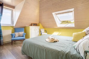 Une chambre avec un plafond en pente, un lit recouvert d’une couverture verte, une chaise bleue, une armoire blanche et une fenêtre donnant sur un paysage verdoyant.