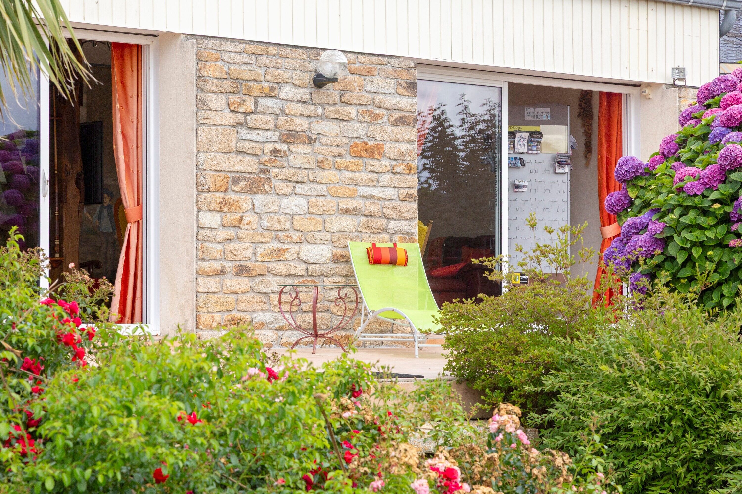 A stone building with a glass door, a green chair outside, and a garden with various plants and flowers.