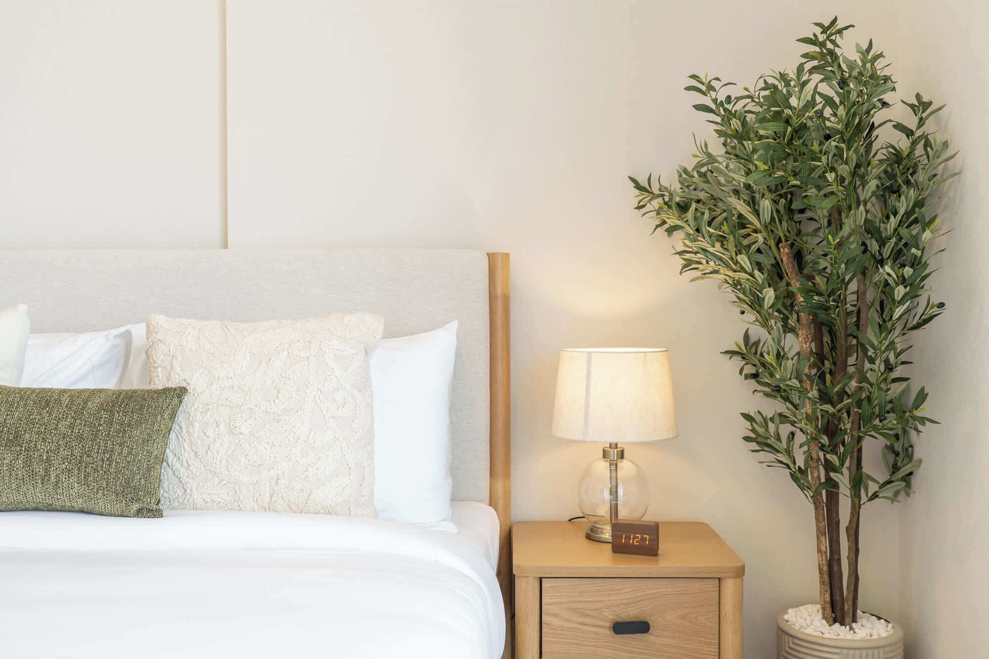 Una cama bien hecha con almohadas blancas y verdes, una lámpara de noche y una planta en maceta.