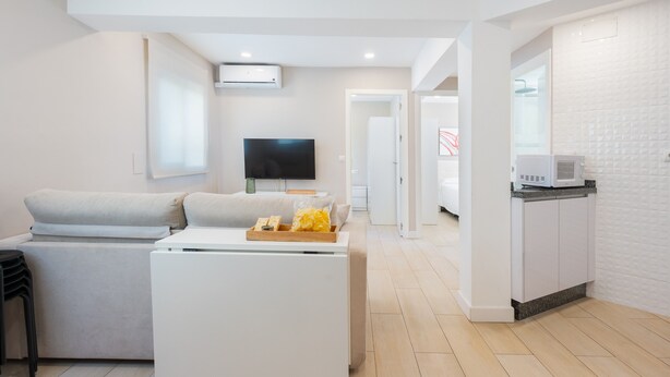A modern living room with a sofa, a flat-screen TV, a white coffee table, and a kitchen area with a microwave.