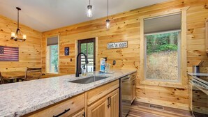 Une cuisine rustique aménagée dans une cabane en bois, avec des comptoirs en granit, un évier, un lave-vaisselle et une grande fenêtre donnant sur une forêt.