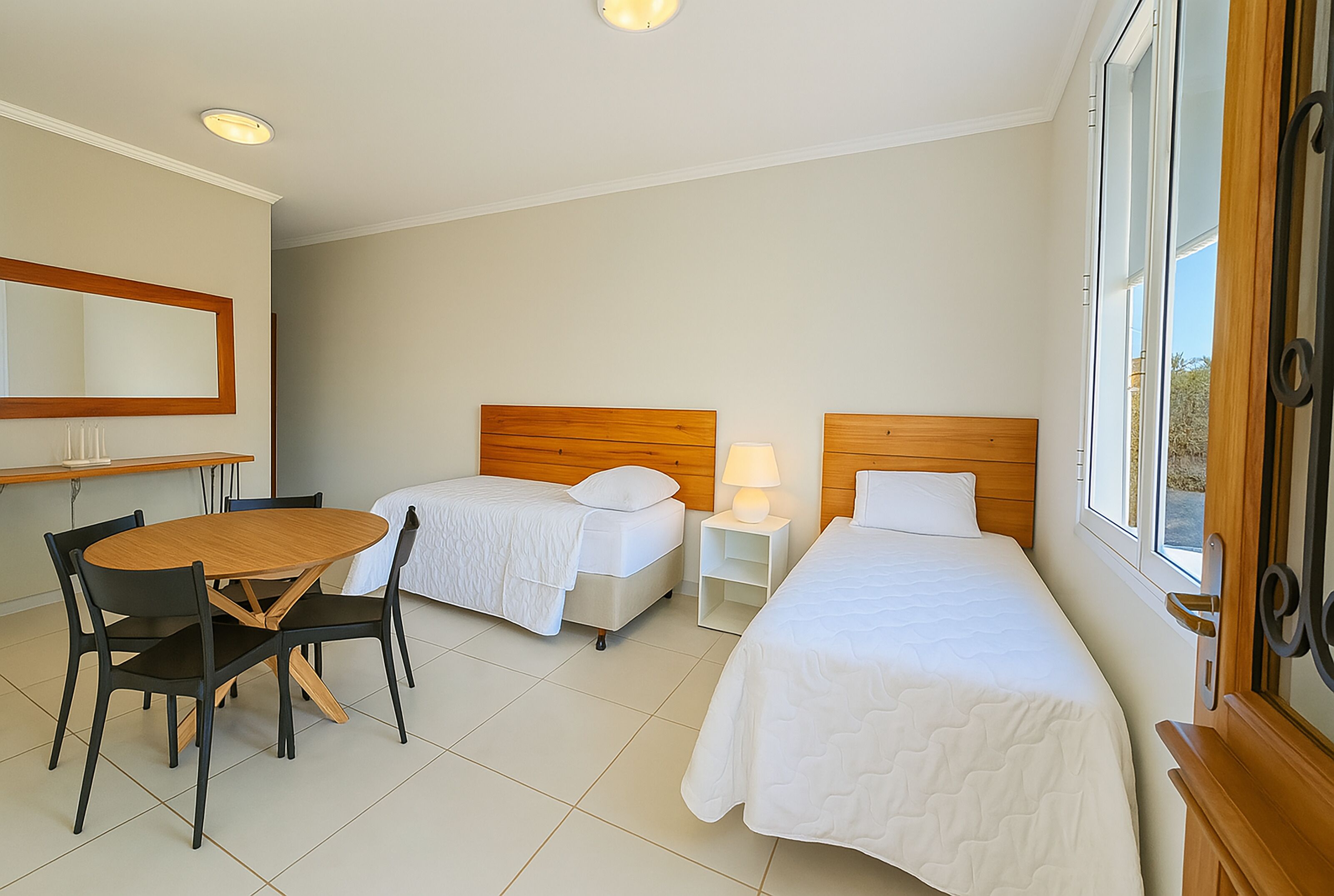 A hotel room with two single beds, a wooden table with chairs, a small bedside table with a lamp, and a large window with a view of the outdoors.