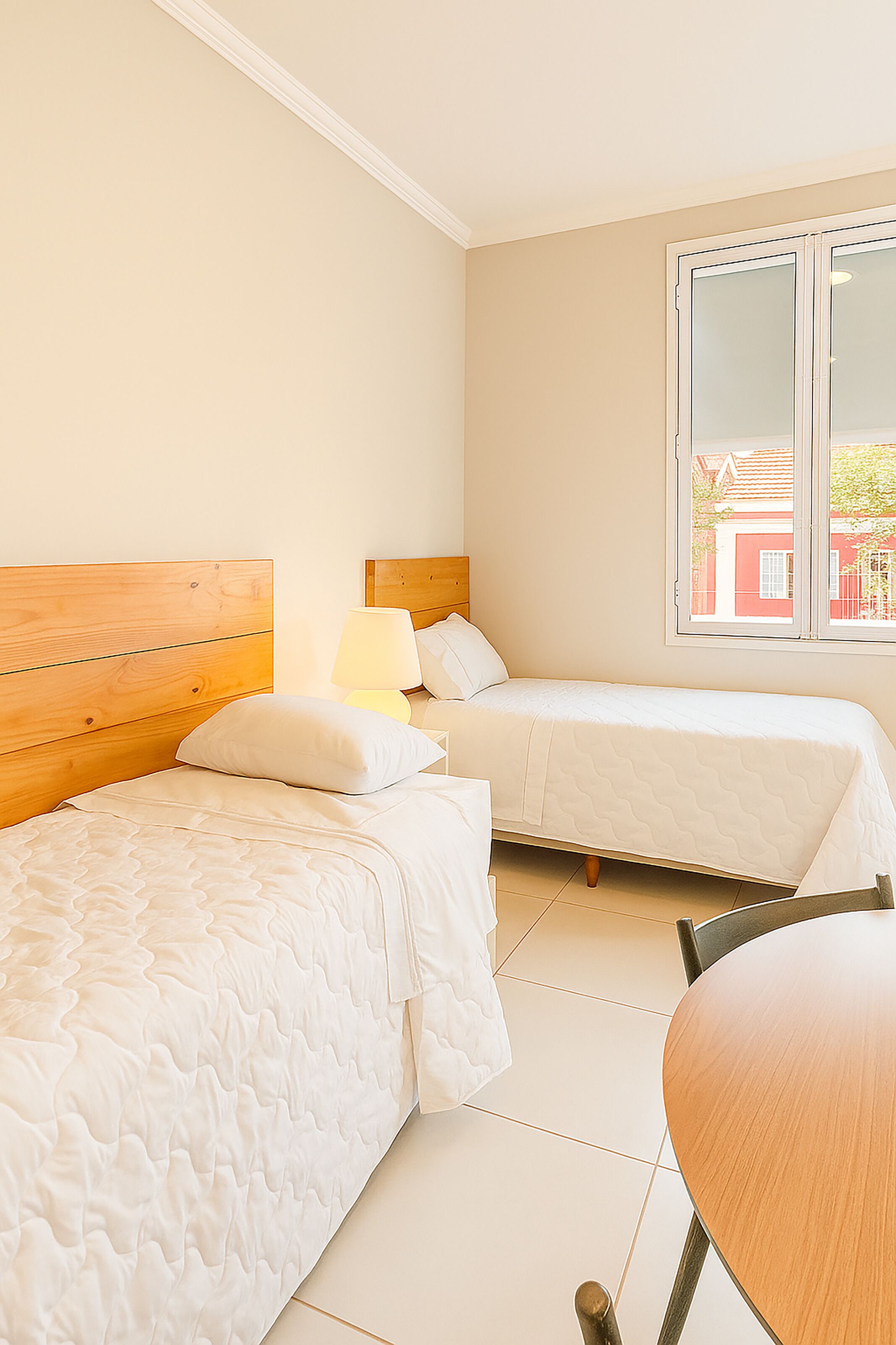 Een slaapkamer met twee bedden, een houten hoofdbord, een nachtkastje met een lamp en een raam met uitzicht op gebouwen.
