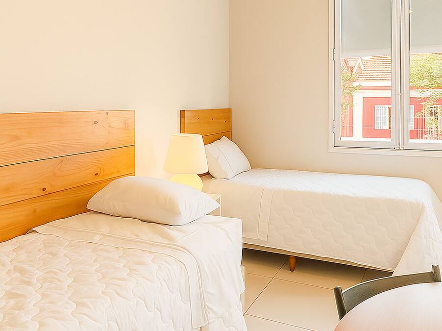 Een slaapkamer met twee bedden, een houten hoofdbord, een nachtkastje met een lamp en een raam met uitzicht op gebouwen.