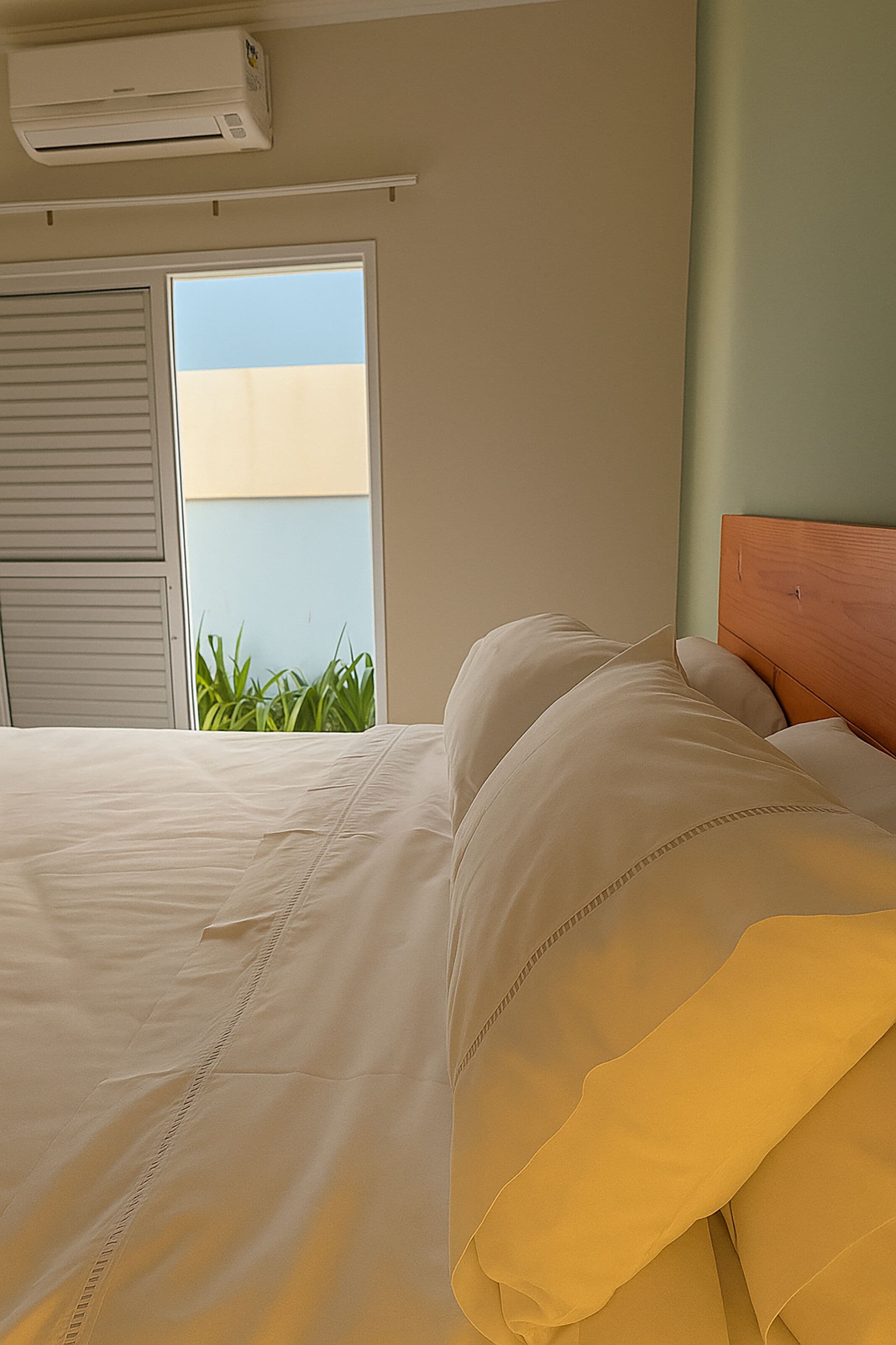 Un dormitorio con cama, cabecera de madera, ventana con persianas y vista a zonas verdes.