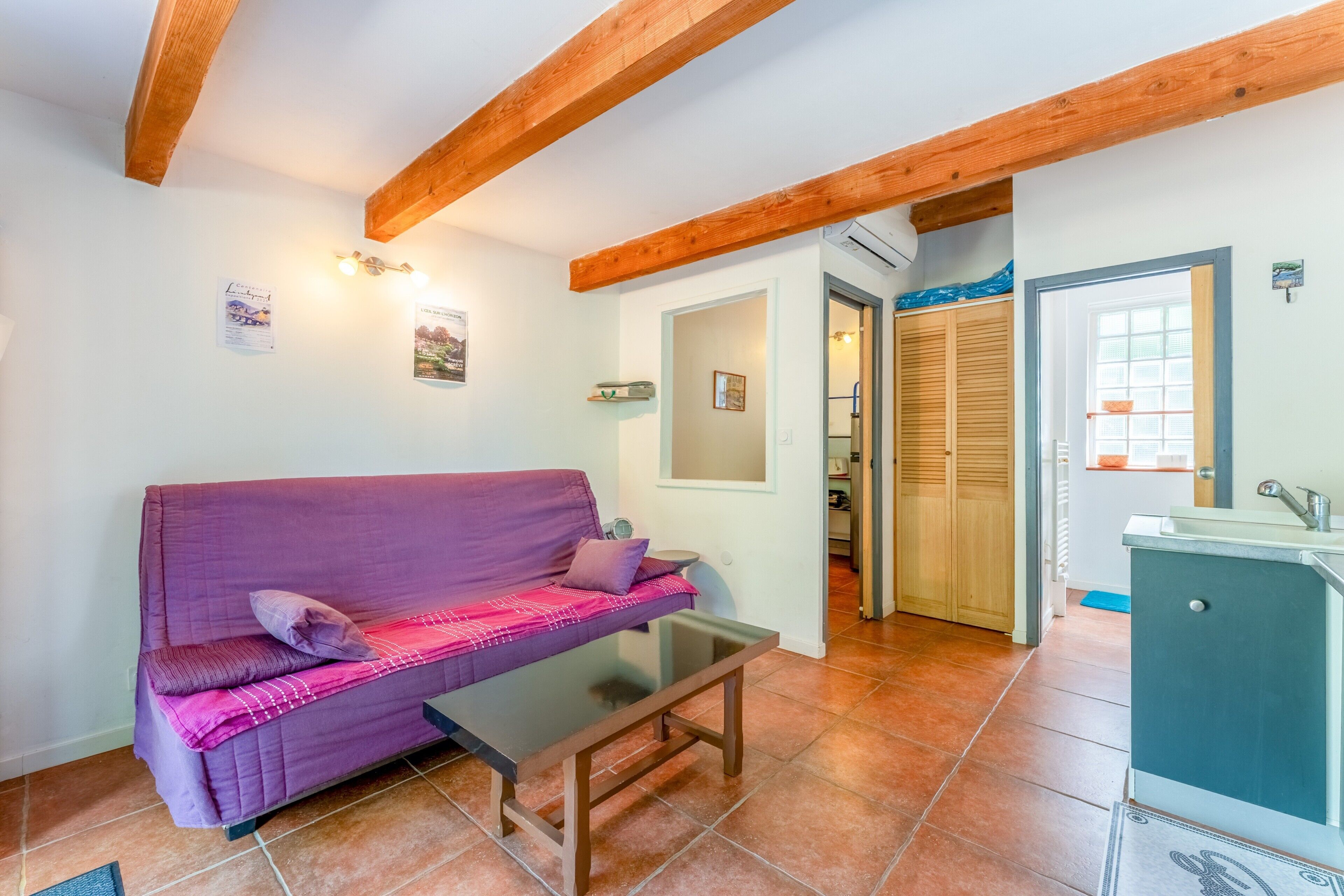 A room with a purple sofa, a small table, wooden beams on the ceiling, a kitchen area, and a bathroom with wooden doors.