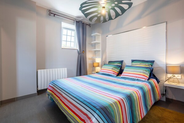 Un dormitorio con una colcha a rayas, una ventana con cortinas, un radiador y una estantería con una lámpara.