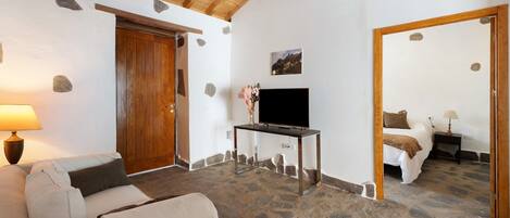 Ein Zimmer mit Steinboden, Holzbalken an der Decke, einem Tisch mit Fernseher, einer Couch und einem Bett mit Nachtisch.