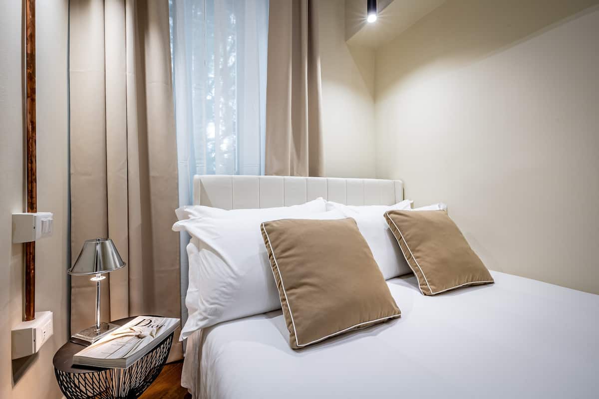 A neatly made bed with white and beige pillows, a bedside lamp, and a book on a bedside table.