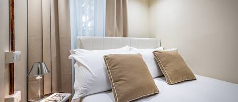 Una cama bien hecha con almohadas blancas y color crema, una lámpara de noche y un libro en una mesita.