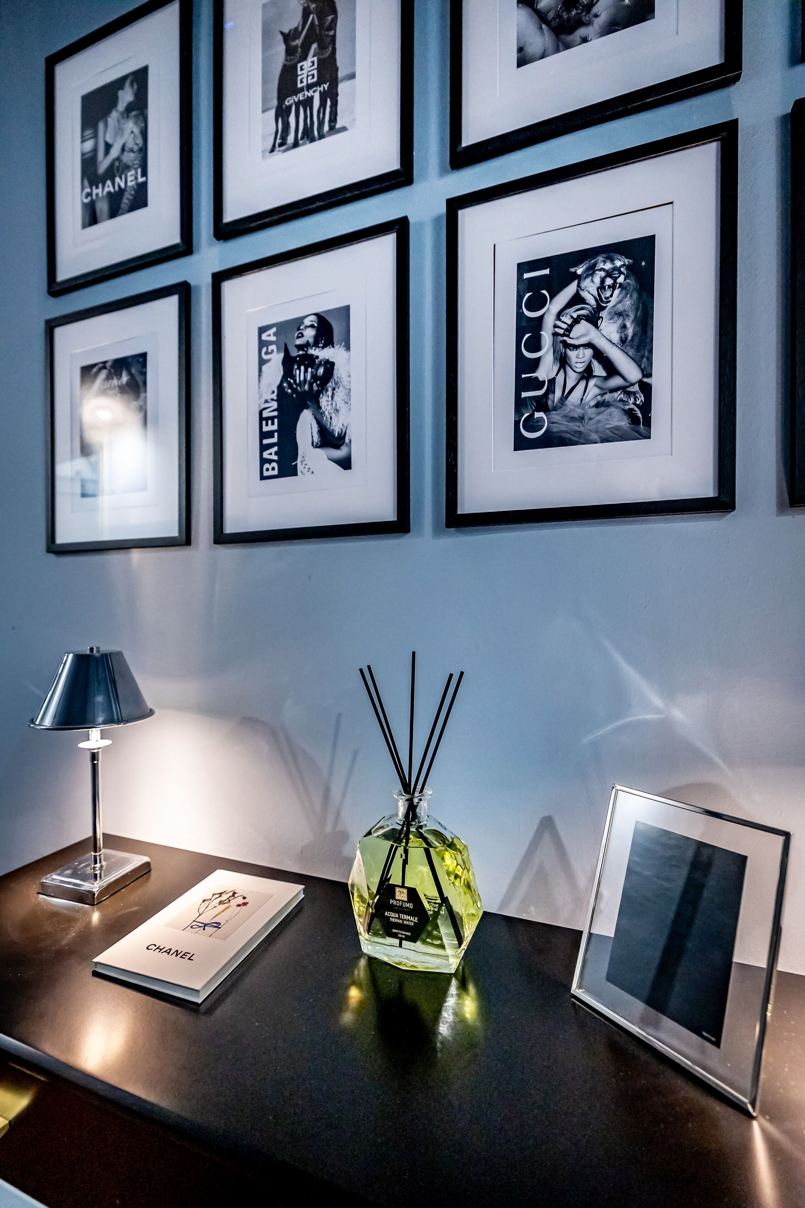 A wall with framed pictures of luxury brands, a table with a lamp, a perfume bottle, and a framed picture.