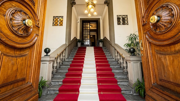 Een brede, statige trap met rode en witte lopers, bekleed met houten deuren met ingewikkelde houtsnijwerken, die leidt naar een kamer met een kroonluchter.