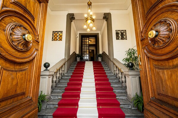 A grand staircase with red and white carpeted steps, flanked by wooden doors with intricate carvings, leading to a room with a chandelier.