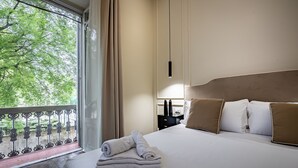 Hotelkamer met een bed, nachtkastje, lamp en uitzicht op bomen door een raam.