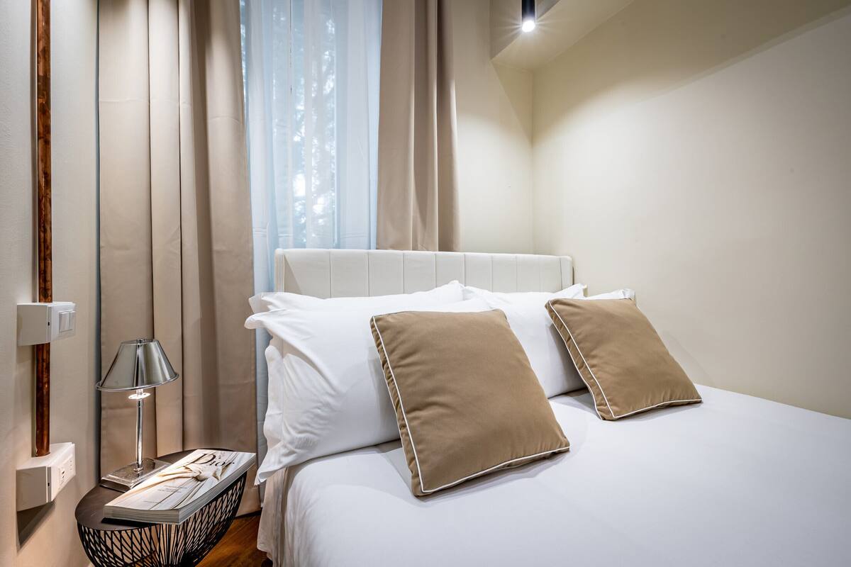 A neatly made bed with white and brown pillows, a bedside lamp, and a book on a bedside table.
