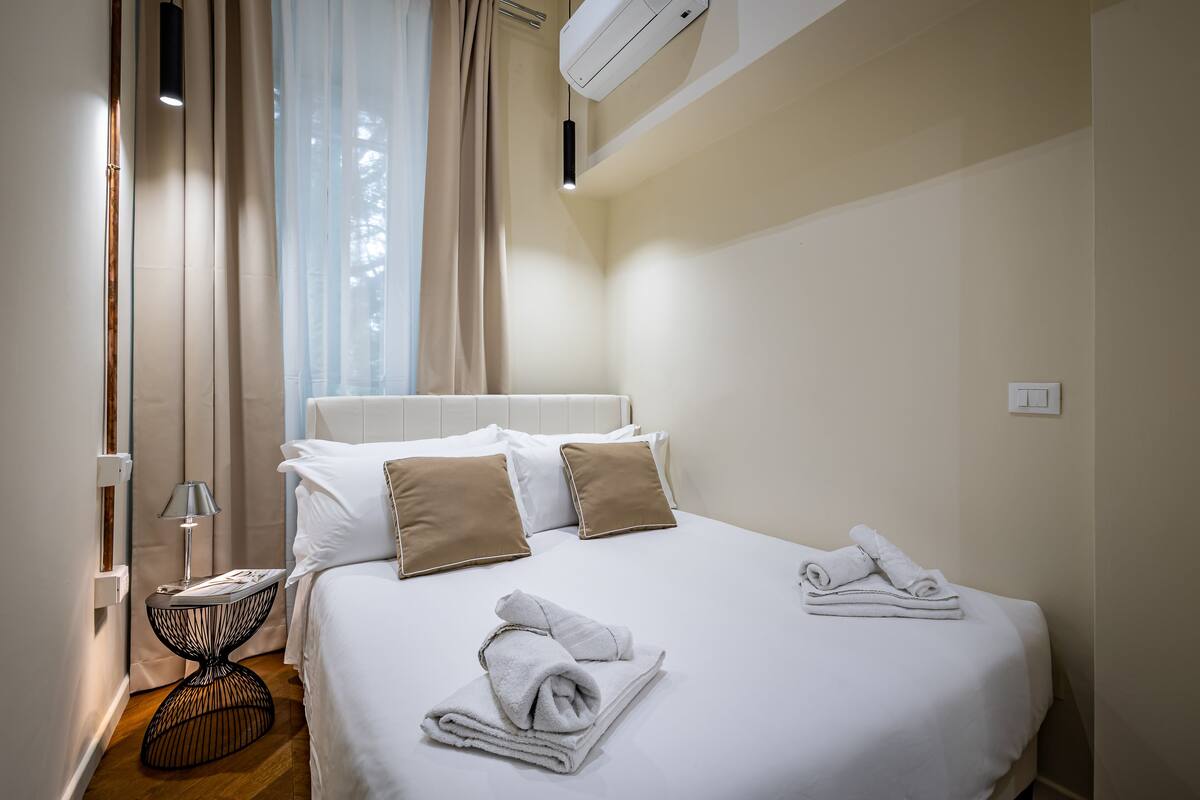 A neatly made bed with white and beige pillows, a bedside table with a lamp, and a window with sheer curtains.