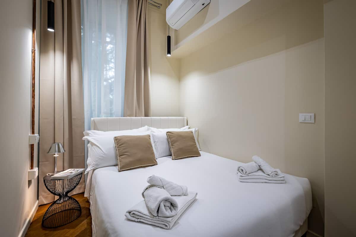 A neatly made bed with white and beige pillows, a bedside table with a lamp, and a window with sheer curtains.