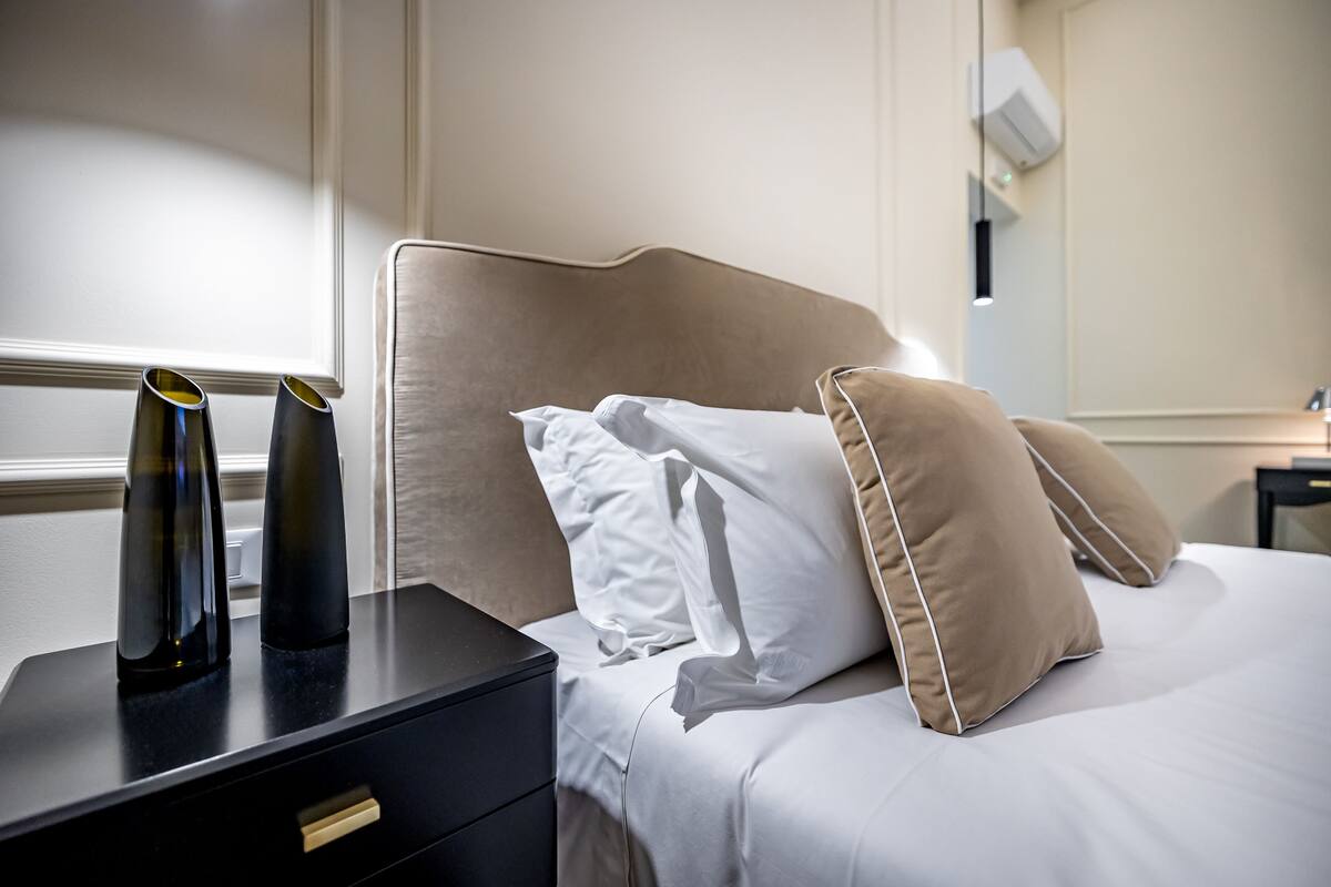 A neatly made bed with white and beige pillows, a black bedside table with two black cylindrical objects, and a wall-mounted air conditioning unit.