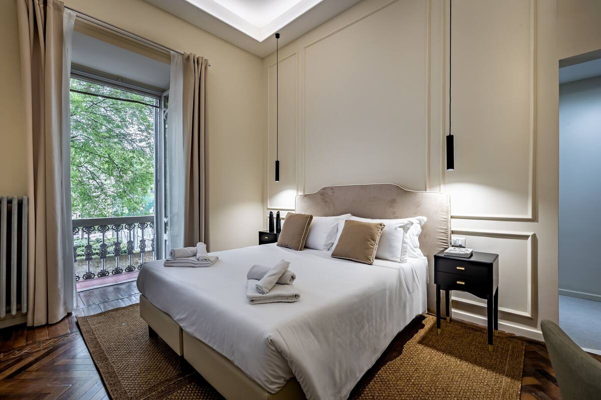 A hotel room with a bed, bedside table, and a view of greenery outside.