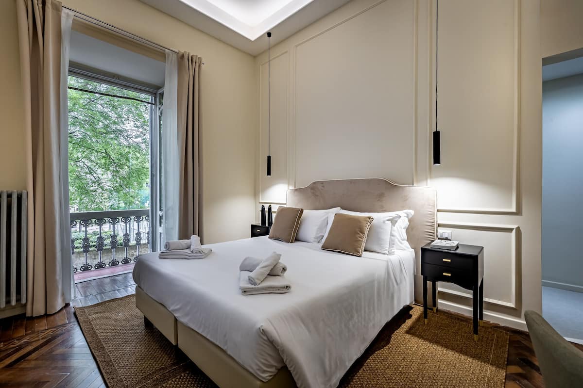 A hotel room with a bed, bedside table, and a view of greenery outside.