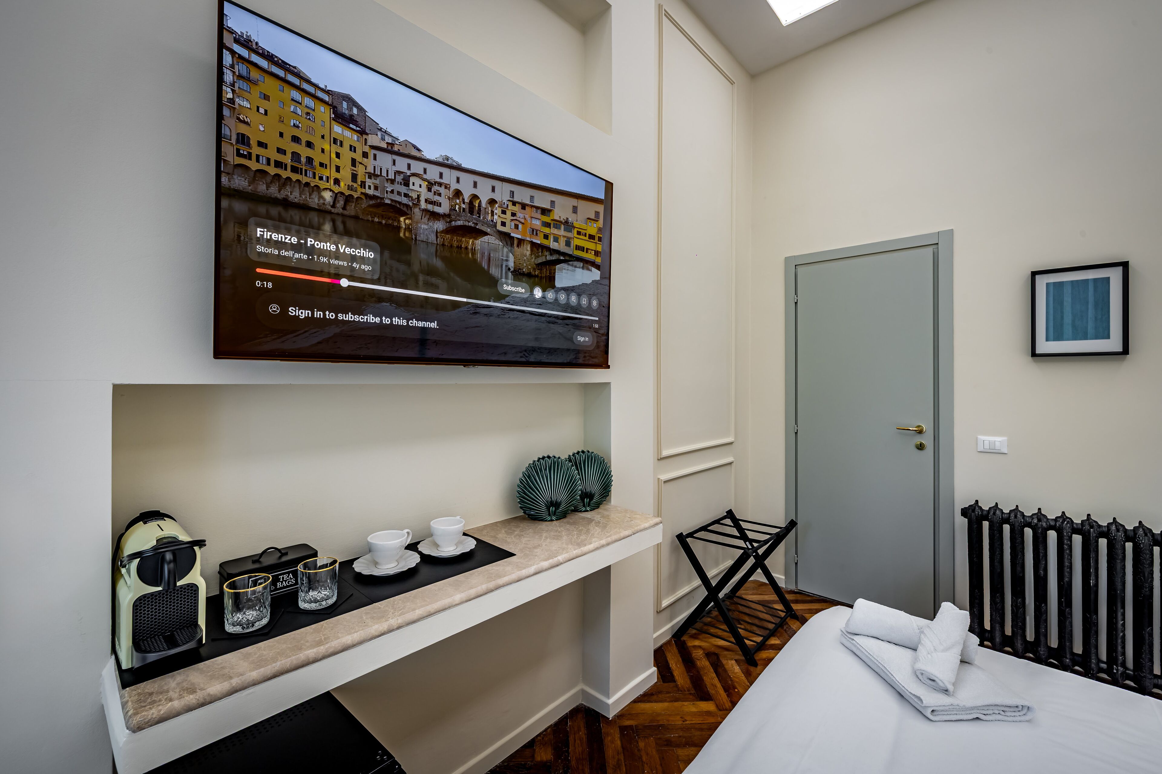 A hotel room with a television displaying a scenic view, a coffee machine, two cups, a tray, and a neatly folded towel on the bed.