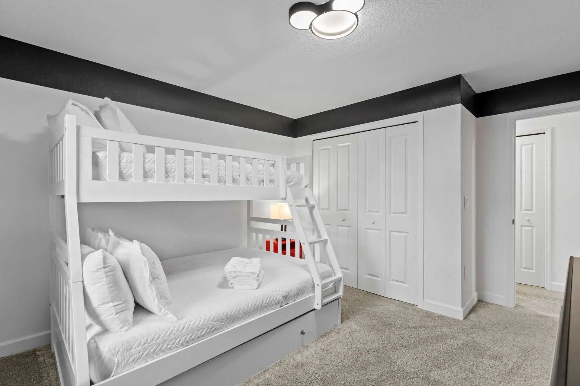 A bunk bed with a ladder, white bedding, and a nightstand with a lamp. There are white doors and a carpeted floor.