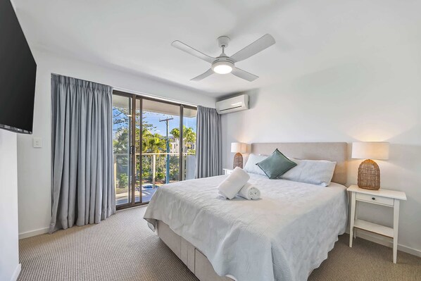 A hotel room with a bed, a ceiling fan, an air conditioning unit, a nightstand with a lamp, and a view of the outdoors through a large window. - Lahaina 9 Beach Access 60 Seconds (Mooloolaba)