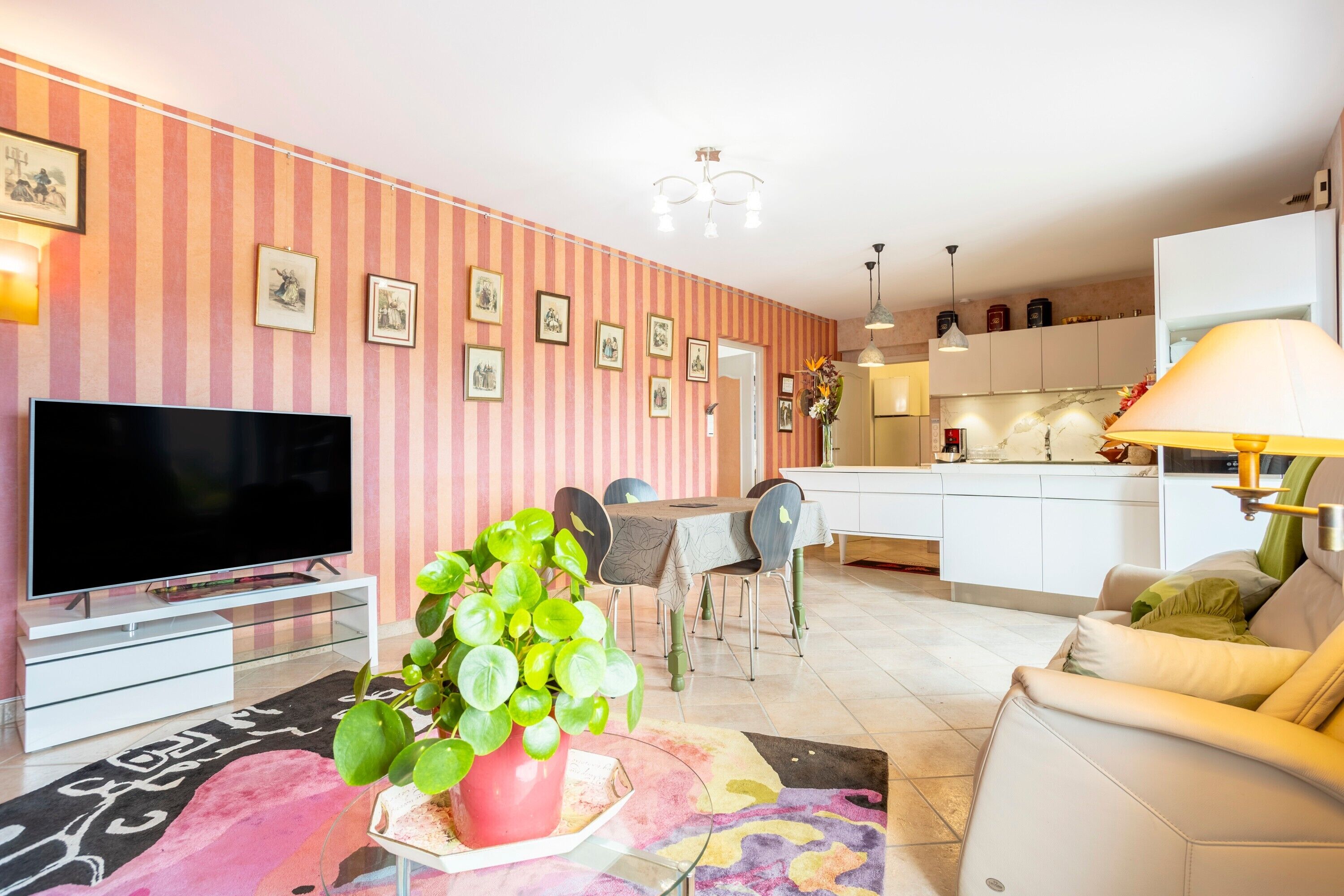 Un salon doté d’un téléviseur à écran plat, d’un canapé, d’un coin repas avec des chaises, d’une cuisine à l’arrière-plan et d’un mur à rayures orné de tableaux encadrés.