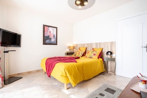 A bedroom with a bed, a television, a framed picture, a bedside table with a lamp, and a door.