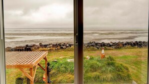 On aperçoit, à travers une fenêtre, une table et des chaises en bois sur une pelouse, avec un rivage rocheux et un phare au loin.