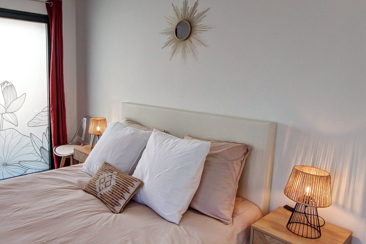 Ein Schlafzimmer mit einem Bett, zwei Kissen, einem Nachttisch mit Lampe und einem Wandspiegel.