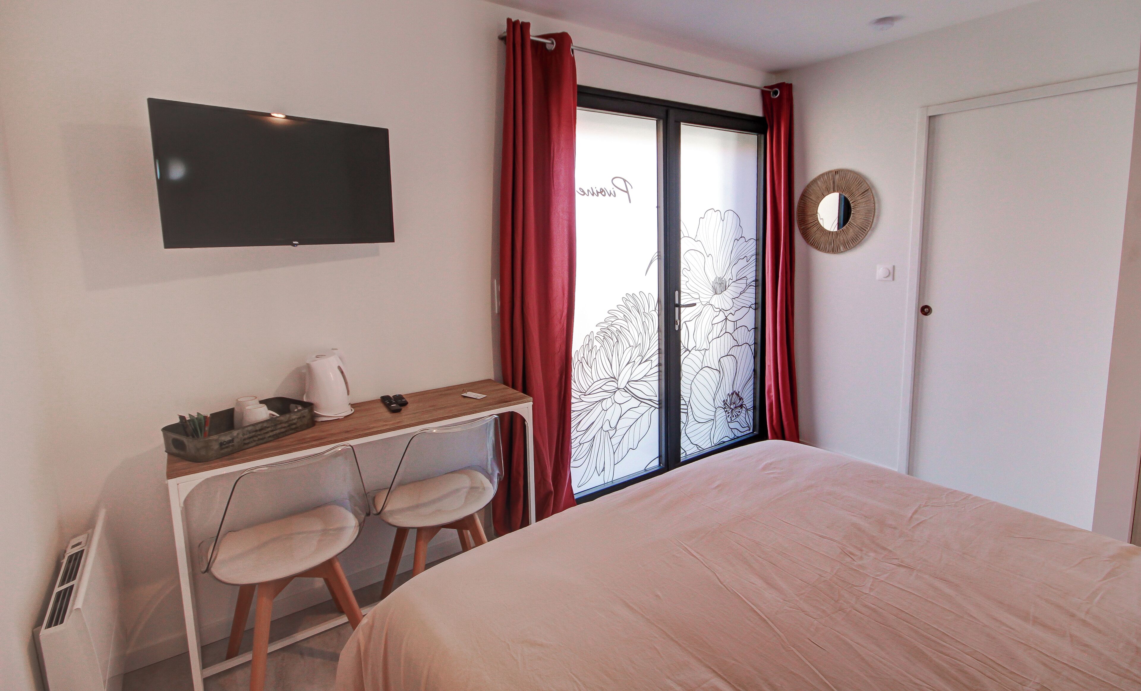 A bedroom with a bed, a desk with two chairs, a television, and a window with red curtains.