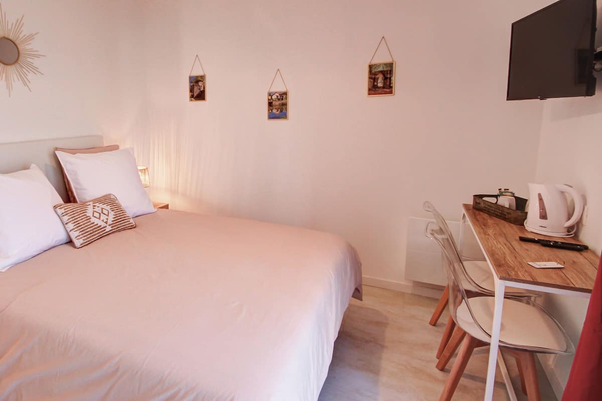 Ein ordentlich bezogenes Bett mit weißer Bettwäsche und dekorativen Kissen, ein Nachttisch mit Lampe, ein Holztisch mit Wasserkocher und ein an der Wand befestigtes Fernsehgerät.