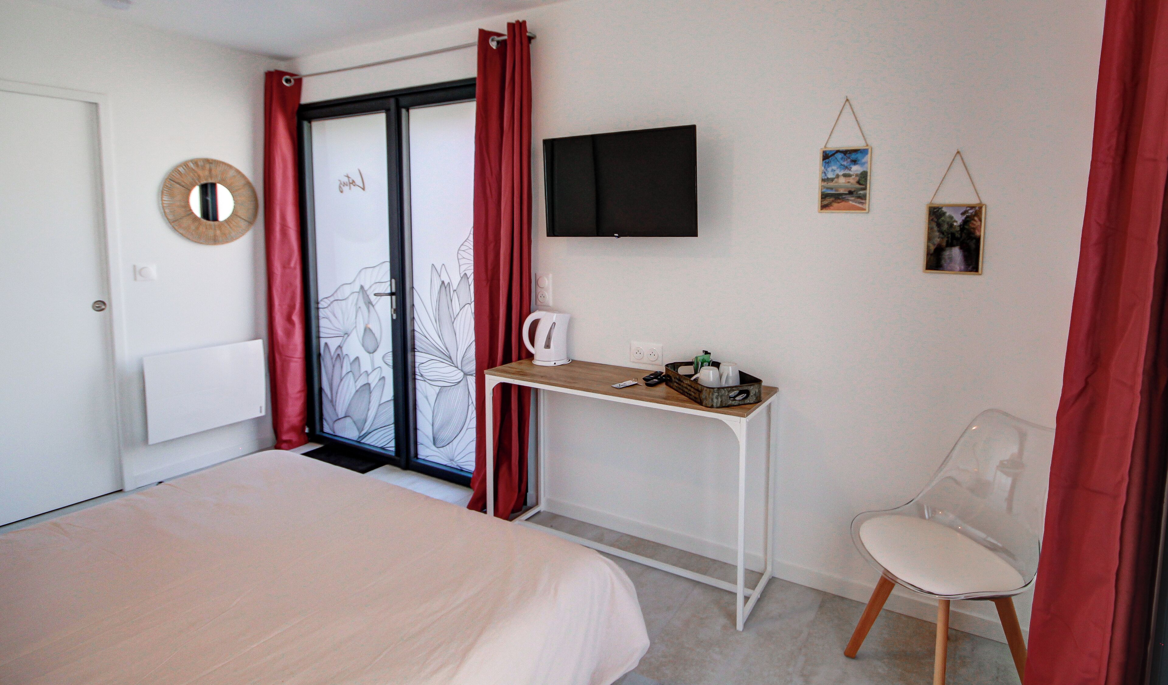 A hotel room with a bed, a desk, a chair, a TV, and a sliding glass door with red curtains.