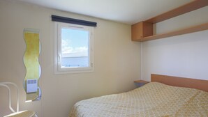 A bedroom with a bed, a chair, a window, and a wall-mounted shelf. - Mobile Home 'Entre Terre Et Mer' with Private Terrace and Private Garden (Coatascorn)