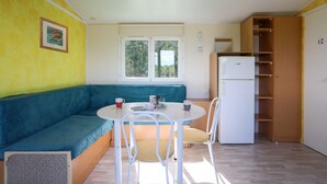 A compact living space with a sofa, a dining table with chairs, a refrigerator, and a window with a view of greenery. - Mobile Home 'Entre Terre Et Mer' with Private Terrace and Private Garden (Coatascorn)