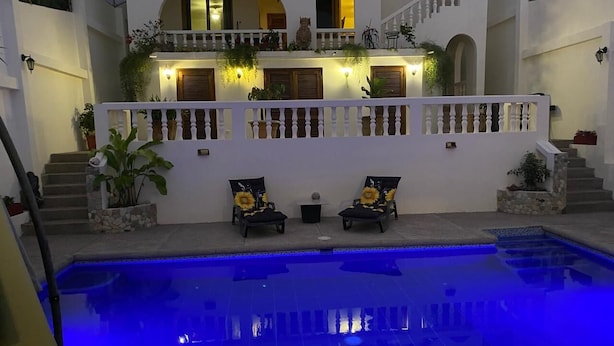 Une piscine dont le bord est illuminé de lumière bleue, deux chaises longues ornées de motifs de tournesols, et un bâtiment blanc doté de balcons et d’escaliers.