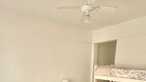 Un dormitori amb un llit individual, una tauleta de nit, una làmpada, un ventilador de sostre i una llitera.