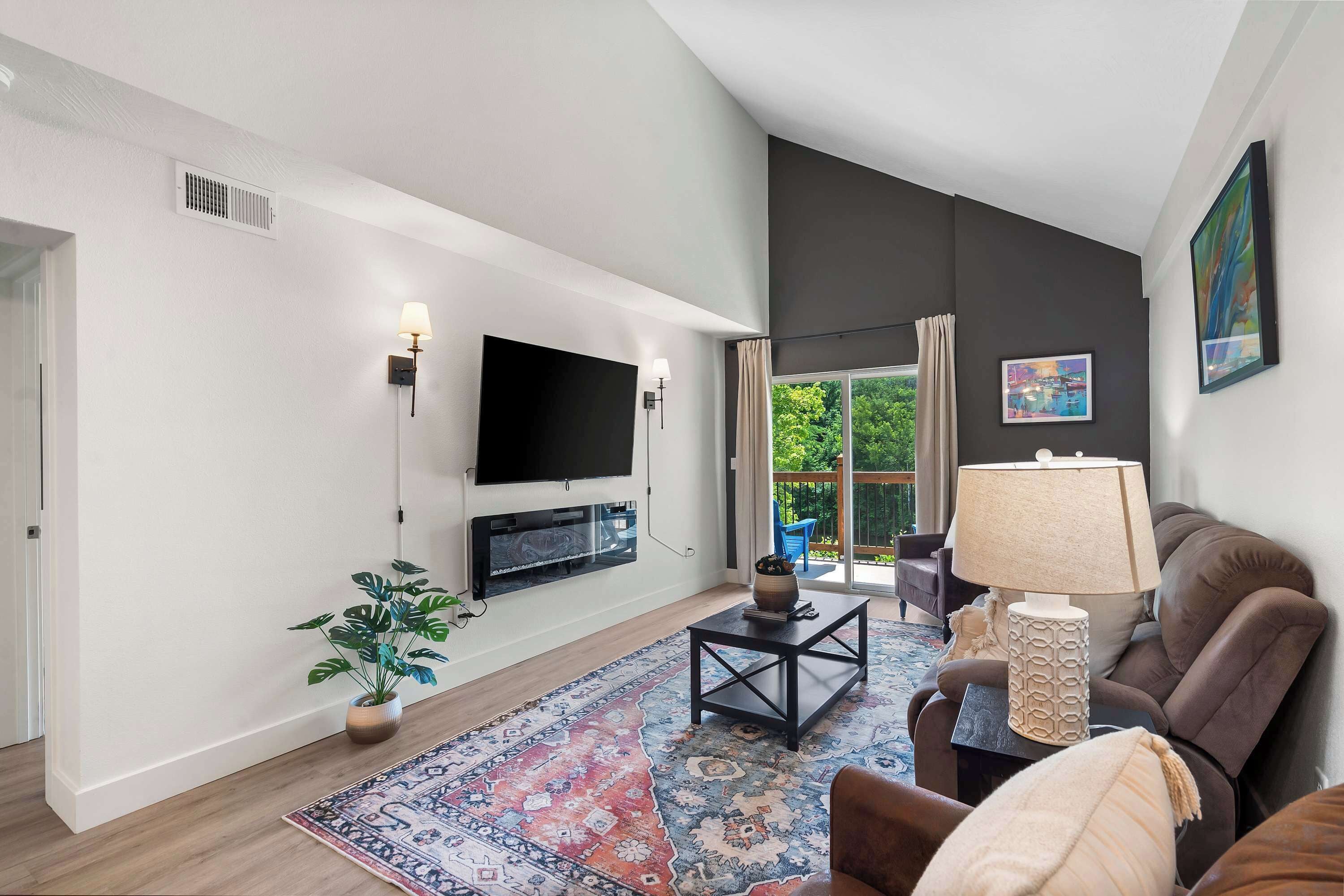 A living room with a flat-screen TV, a sofa, a coffee table, a patterned rug, and a potted plant.